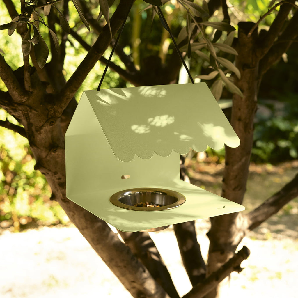 A green Picoti bird feeder with a stainless steel bowl, hanging from a cord in a garden setting.