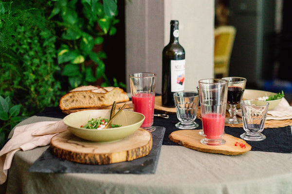 Table setting with a bowl of salad, glasses of pink and red drinks, and a bottle of wine outdoors.