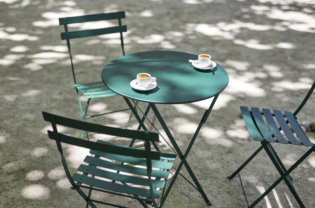 A green folding Bistro table with two cups on top, accompanied by two chairs, set outdoors with shadows on the ground.