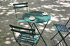 A green folding Bistro table with two cups on top, accompanied by two chairs, set outdoors with shadows on the ground.