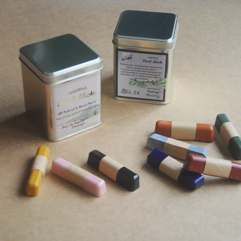 Two metal tin candles with silver lids and cream labels are displayed on a beige surface, alongside several small wooden stick candles in assorted colors including yellow, pink, black, blue, red, and green.
