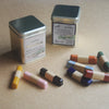 Two metal tin candles with silver lids and cream labels are displayed on a beige surface, alongside several small wooden stick candles in assorted colors including yellow, pink, black, blue, red, and green.