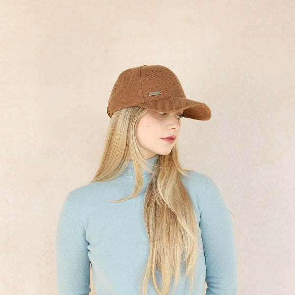 A person wearing a brown, spice-colored, classic baseball style cap with an adjustable strap, against a neutral background.