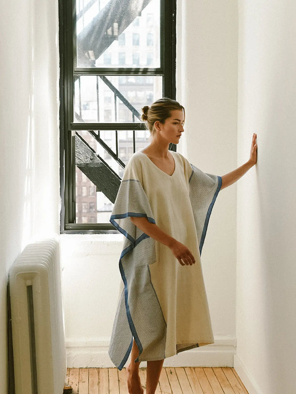 Woman wearing a light-colored dress with blue trim standing in a room with large windows.