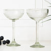 Two clear glass coupe glasses on a white background with grapes and leaves.