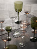 A selection of elegant, mouth-blown glassware including wine glasses, cocktail glasses, and liqueur glasses, displayed on a table. The glassware comes in clear and various colored tints.