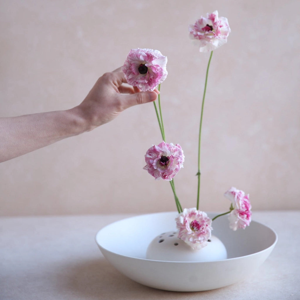 Hand arranging pink flowers in a white vase on a light surface with a neutral background