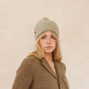 Woman wearing a sage green beanie and brown coat against a beige background