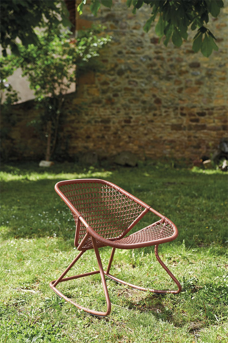 A brown aluminum and fiber woven rocking chair on a grassy outdoor area, with a stone wall in the background.