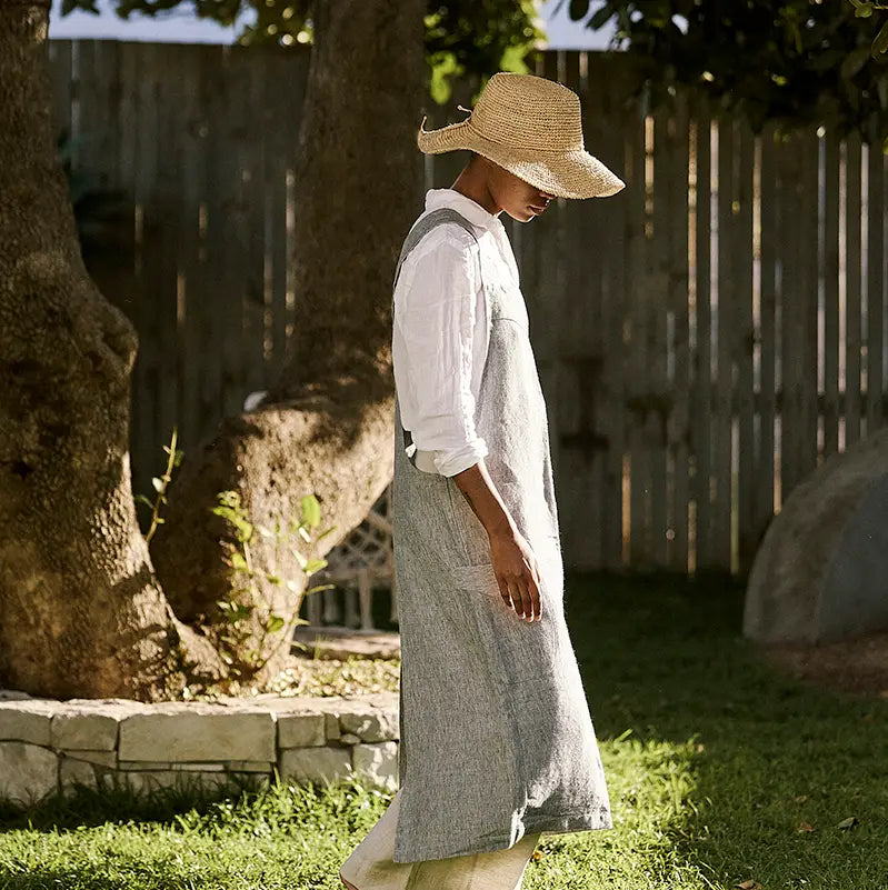 A woman wearing a straw hat, white button-up shirt, and a long gray linen apron walks outdoors in a sunny backyard with a wooden fence and grass.