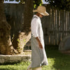 A woman wearing a straw hat, white button-up shirt, and a long gray linen apron walks outdoors in a sunny backyard with a wooden fence and grass.