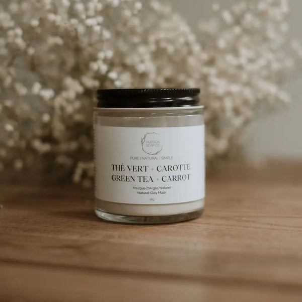 A glass jar containing a natural clay mask labeled 'THE VERB - CAROTTE GREEN TEA + CARROT' placed on a wooden surface with a dry green plant in the background.