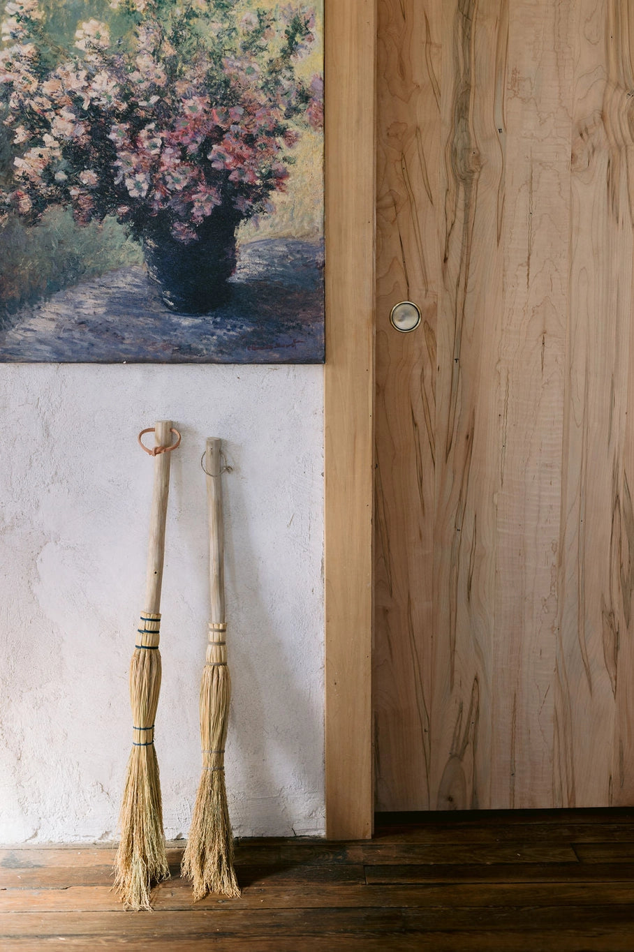 Two brooms leaning against a wooden door with a painting of flowers on the wall.