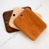 Three rectangular wooden cutting boards with rounded corners and a hole near one end, stacked slightly offset on a white marble surface. The boards show different wood tones: one dark brown, one light beige, and one warm medium brown in the foreground.