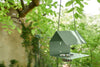 A green Picoti bird feeder with a stainless steel bowl, hanging from a cord in a garden setting.