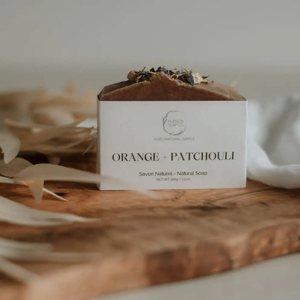 A bar of natural soap with 'ORANGE + PATCHOULI' labeled on the front, placed on a wooden surface with decorative elements around it.