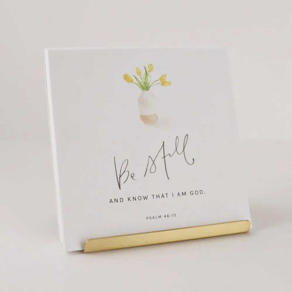 A white square art print or card with a watercolor flower vase, the phrase 'Be Still AND KNOW THAT I AM GOD.' in black script and serif text, and 'PSALM 46:10' at the bottom, displayed on a gold stand against a light neutral background.