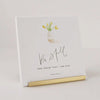 A white square art print or card with a watercolor flower vase, the phrase 'Be Still AND KNOW THAT I AM GOD.' in black script and serif text, and 'PSALM 46:10' at the bottom, displayed on a gold stand against a light neutral background.