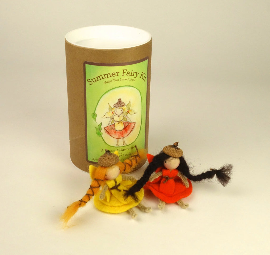 A cylindrical cardboard container labeled 'Summer Fairy Kit' stands behind two small handmade fairy dolls with acorn hats. The dolls wear a yellow dress and an orange dress, with one having long black yarn hair and the other shorter dark hair.