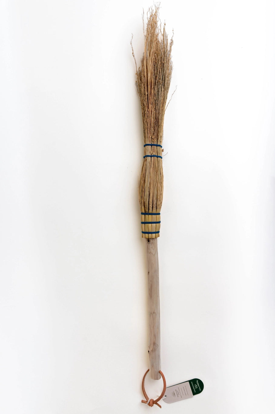 A miniature hand-woven straw broom with a light wood handle and blue wrapping bands, shown against a plain white background with a small tag attached.