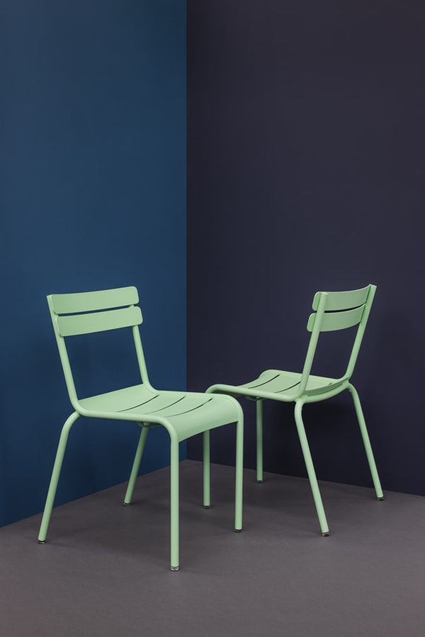 Two green Luxembourg side chairs with tubular aluminum frame and curved aluminum slats, against a dark blue background.
