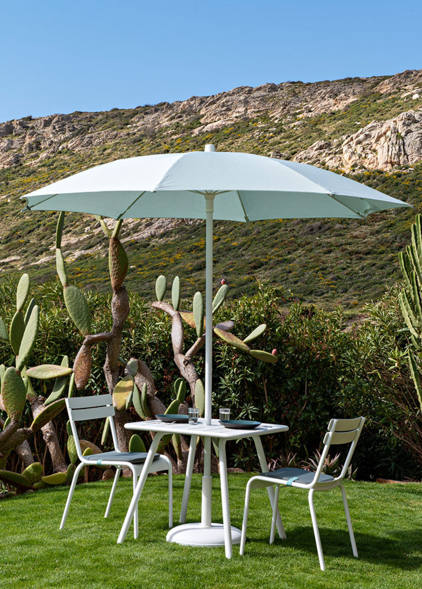 A cotton white Fermob Luxembourg Pedestal Table with two matching chairs with greenery in the background.