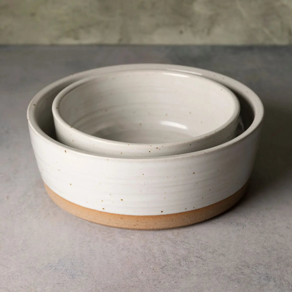 A set of two nested ceramic bowls with a speckled off-white glaze and a natural tan clay base, photographed on a textured gray surface.