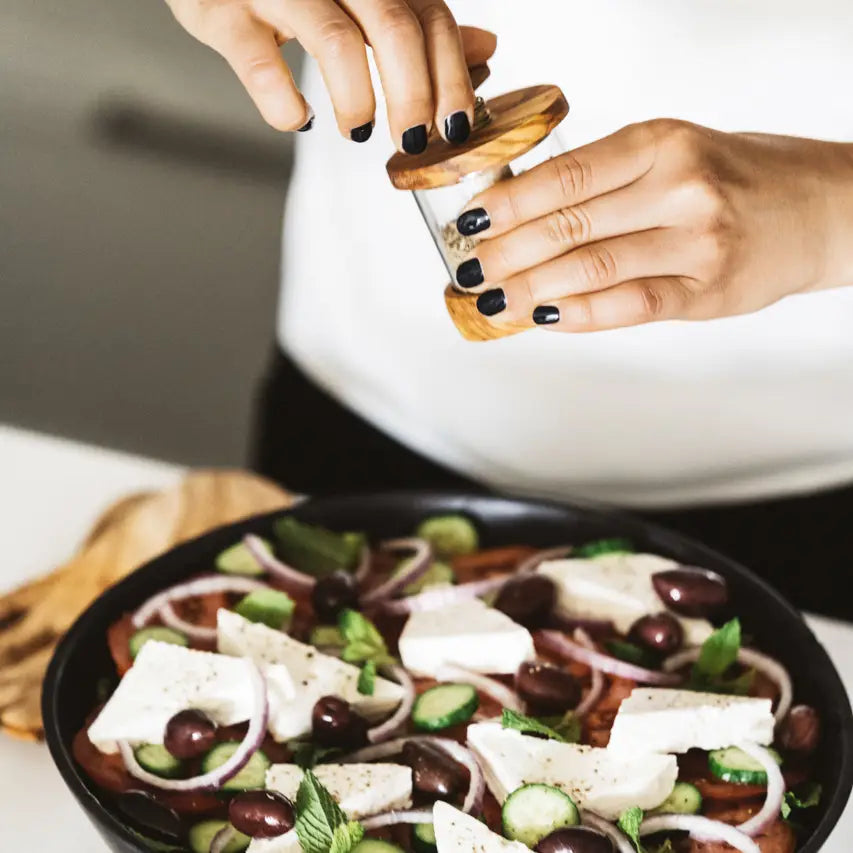 A person's hands using an antique olive wood soft spices grinder over a dish of food, predominantly featuring olives and cheese.