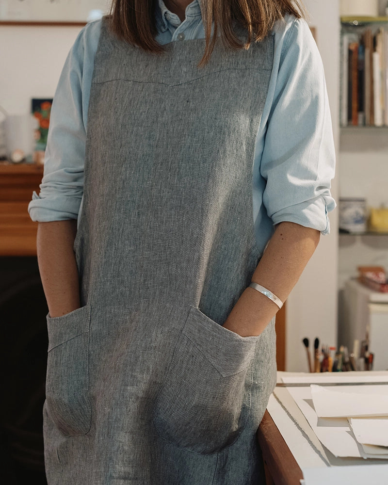 A person wearing a light blue button-up shirt and a gray linen apron with large front pockets, standing in a softly lit indoor room with shelves and desk items in the background.