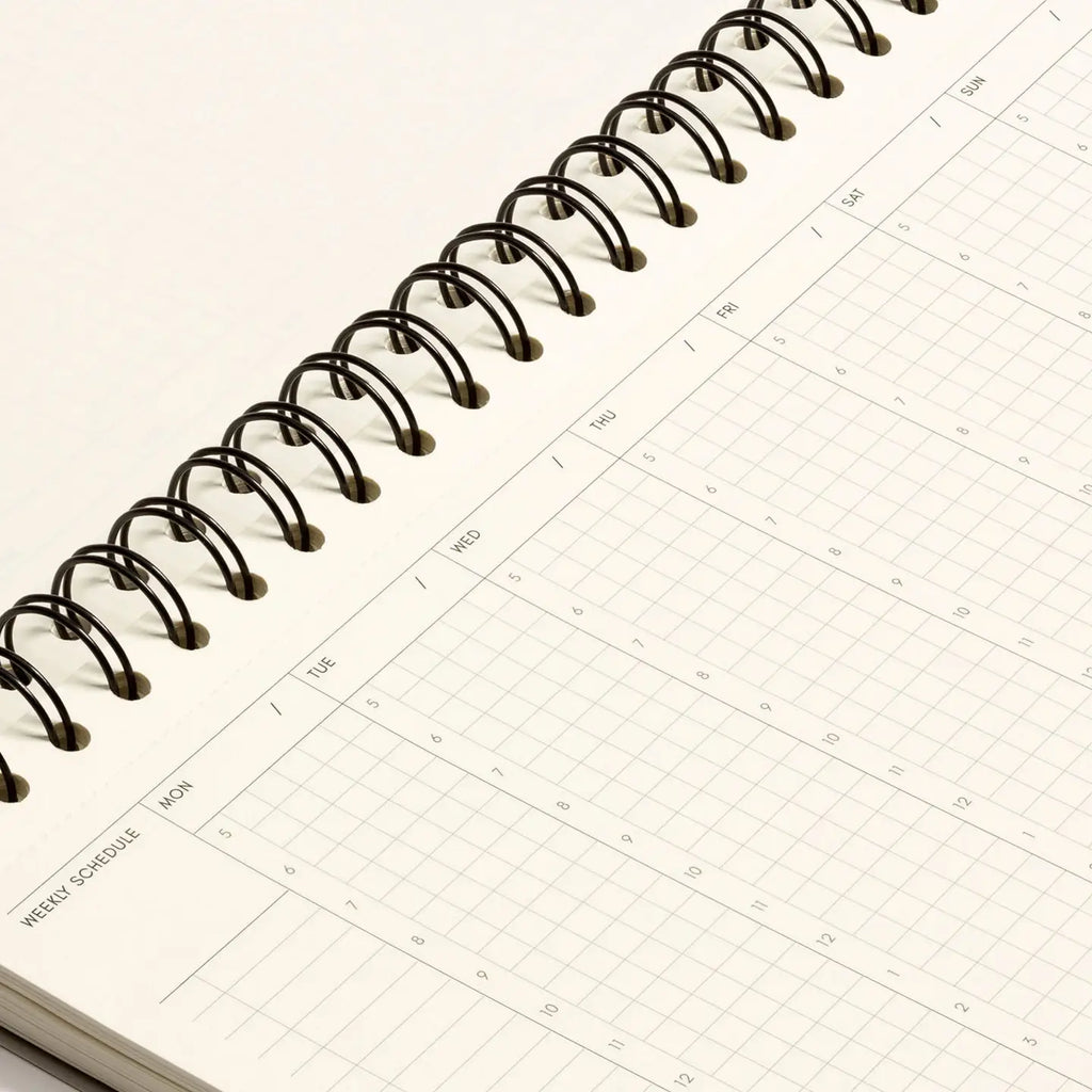 A close-up of an open spiral-bound planner or calendar notebook showing a weekly schedule page with a black wire coil binding on a light neutral background.