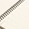A close-up of an open spiral-bound planner or calendar notebook showing a weekly schedule page with a black wire coil binding on a light neutral background.