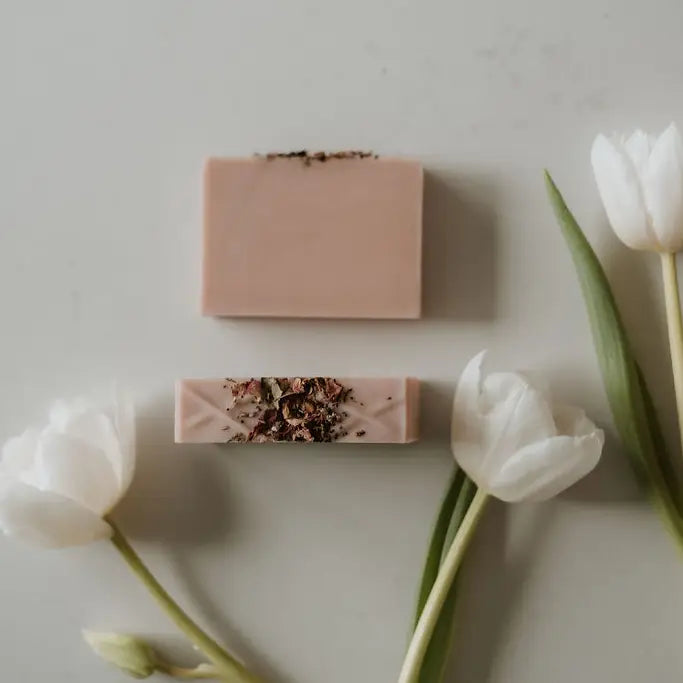 A bar of natural soap placed on a wooden surface with decorative elements around it. White tulips laying around the soap