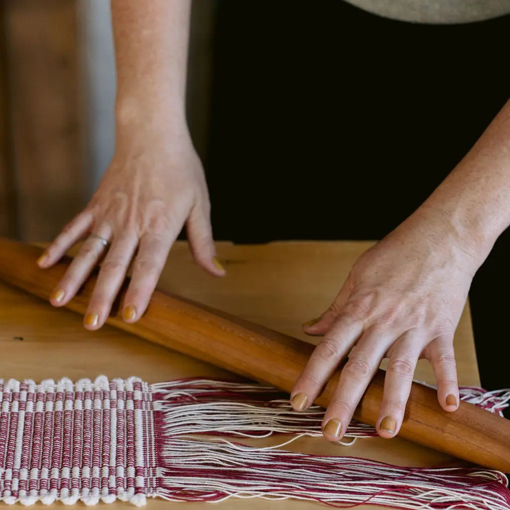 Close-up of a person's hands with weaving textile with burgundy and white threads over a wooden loom, with a wooden rolling pin resting across the woven area.