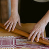 Close-up of a person's hands with weaving textile with burgundy and white threads over a wooden loom, with a wooden rolling pin resting across the woven area.