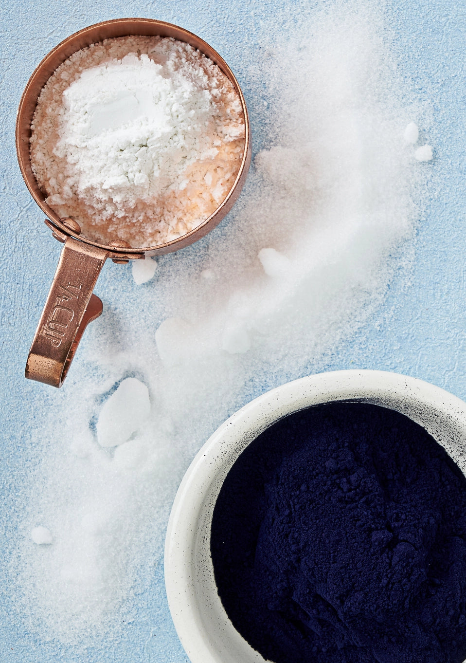 Copper measuring cup with white powder next to a bowl of dark powder on a light blue background