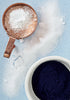 Copper measuring cup with white powder next to a bowl of dark powder on a light blue background