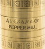 Close-up of a cylindrical brass or gold-toned metal object engraved with the words 'ALEXANDER PEPPER MILL' and decorated with floral motifs.