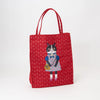 A red canvas tote bag with a repeating floral pattern and a central illustration of a black-and-white cat wearing a blue and pink dress, standing on a white background.