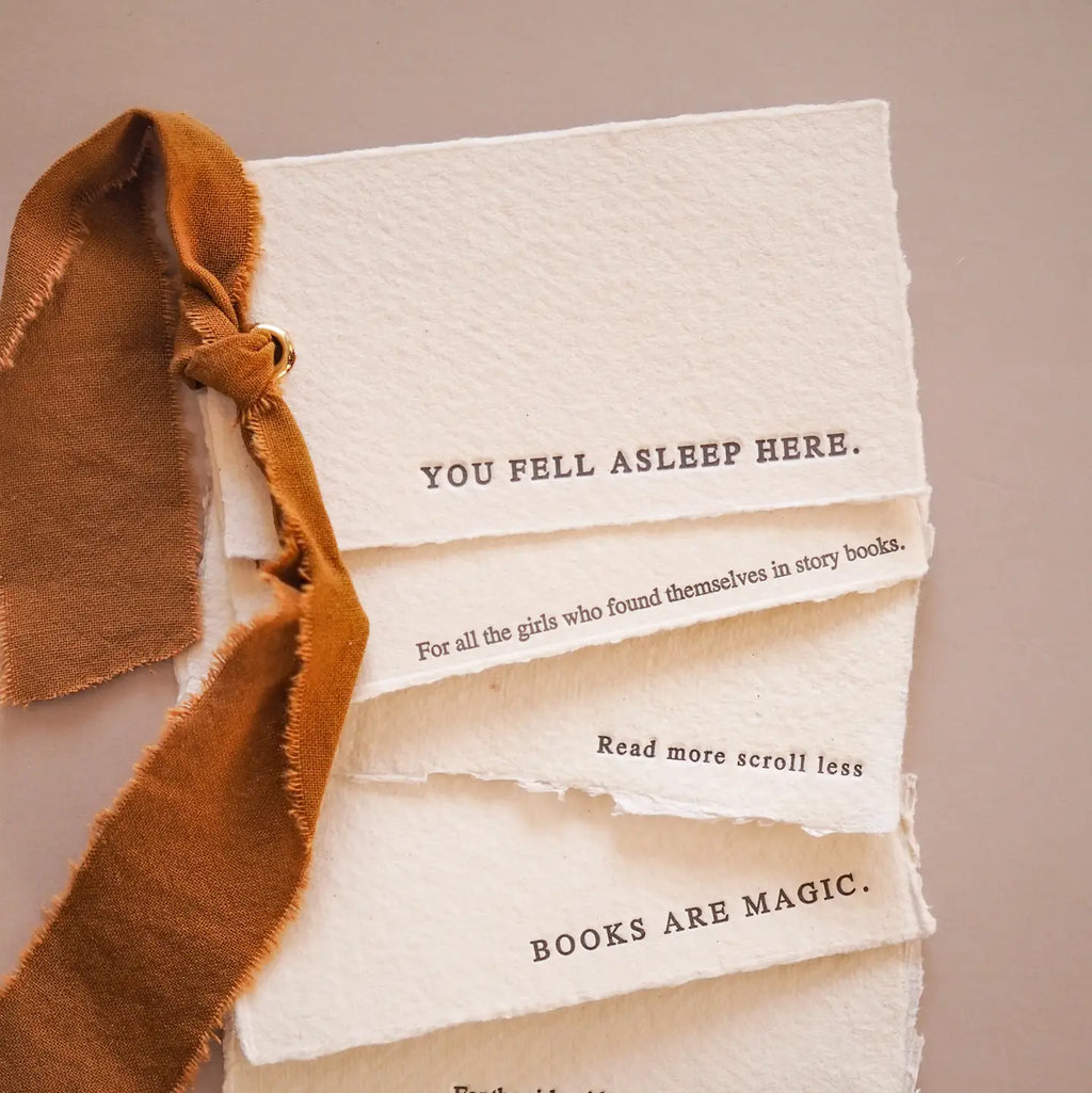 A close-up of a handmade layered off-white paper pages featuring printed quotes, tied with a rustic brown suede ribbon, on a neutral beige background.