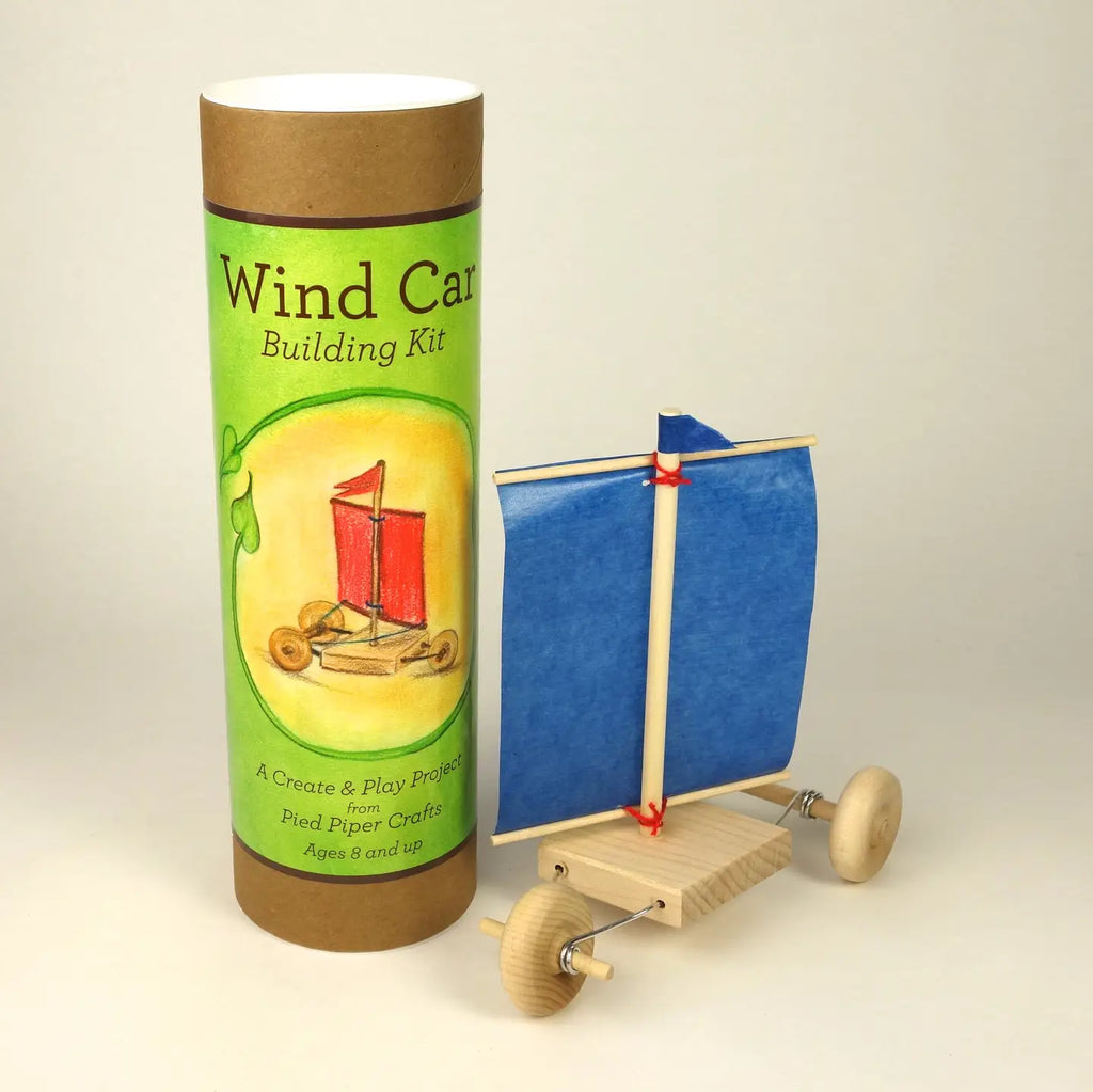 A wind car building kit shown with its green and brown cardboard packaging tube next to the assembled wooden toy wind car with a blue paper sail on a light studio background.