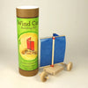 A wind car building kit shown with its green and brown cardboard packaging tube next to the assembled wooden toy wind car with a blue paper sail on a light studio background.