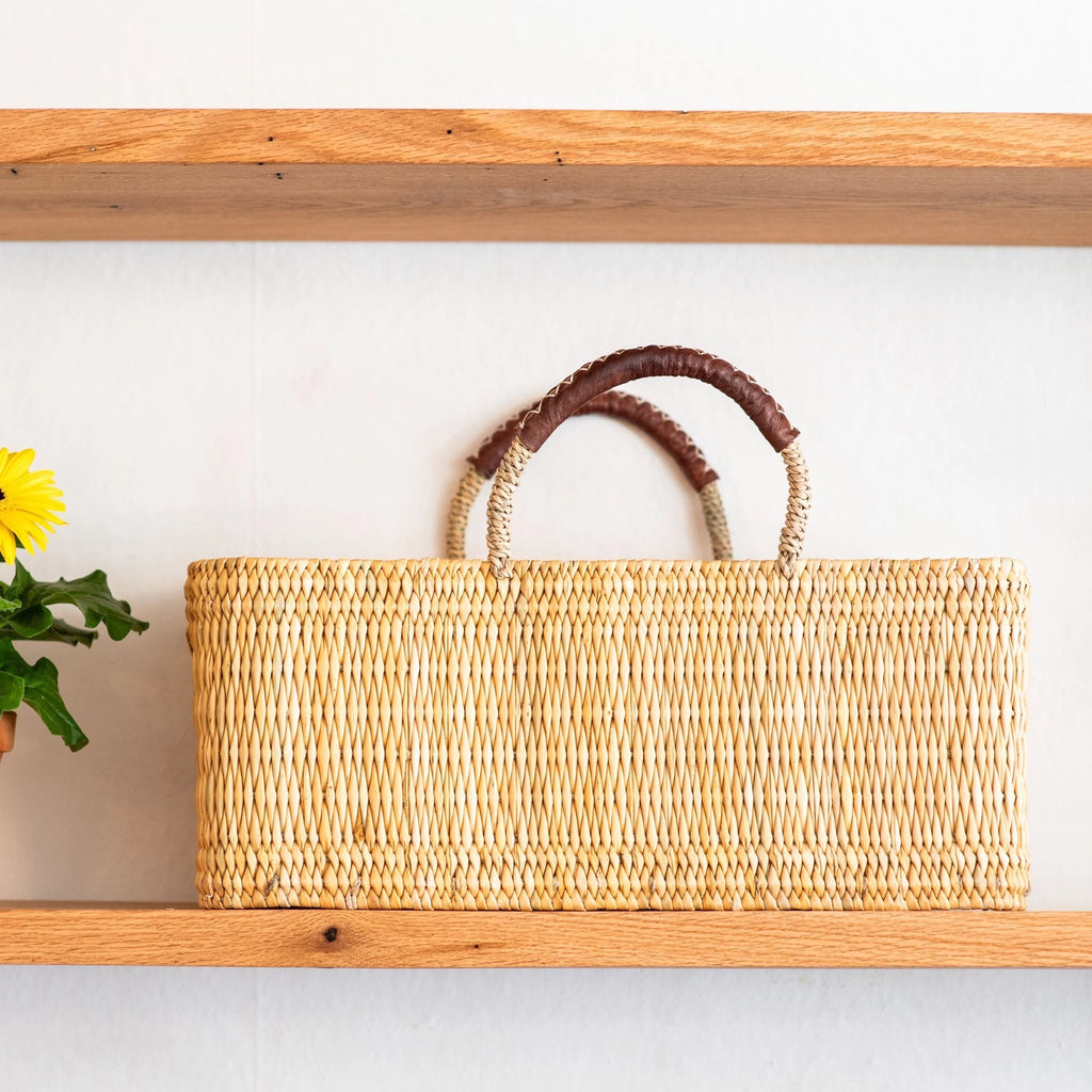 Woven basket with brown handles on a wooden shelf against a white wall.
