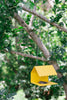 Yellow bird feeder hanging from a tree branch with green foliage in the background
