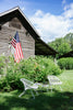 Outdoor setting with a wooden cabin, American flag, and metal chairs on a grassy area.