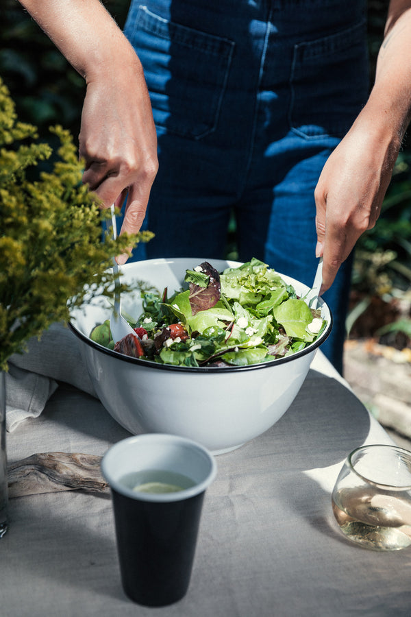 Person preparing a salad in a large white bowl with a cup of dressing on a table.