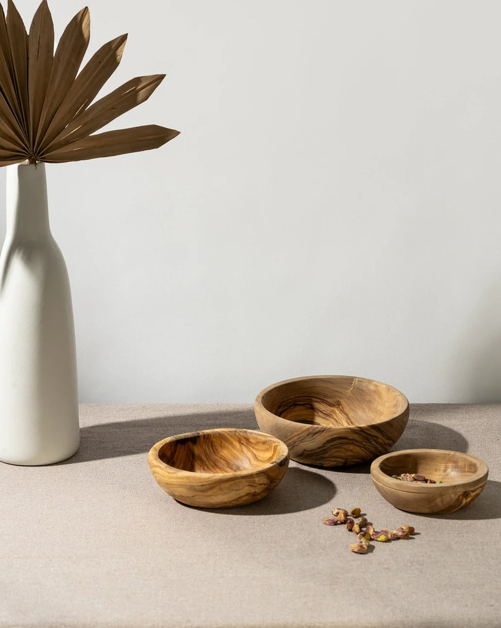 Three wooden bowls on a neutral surface with a white vase and dried leaves.