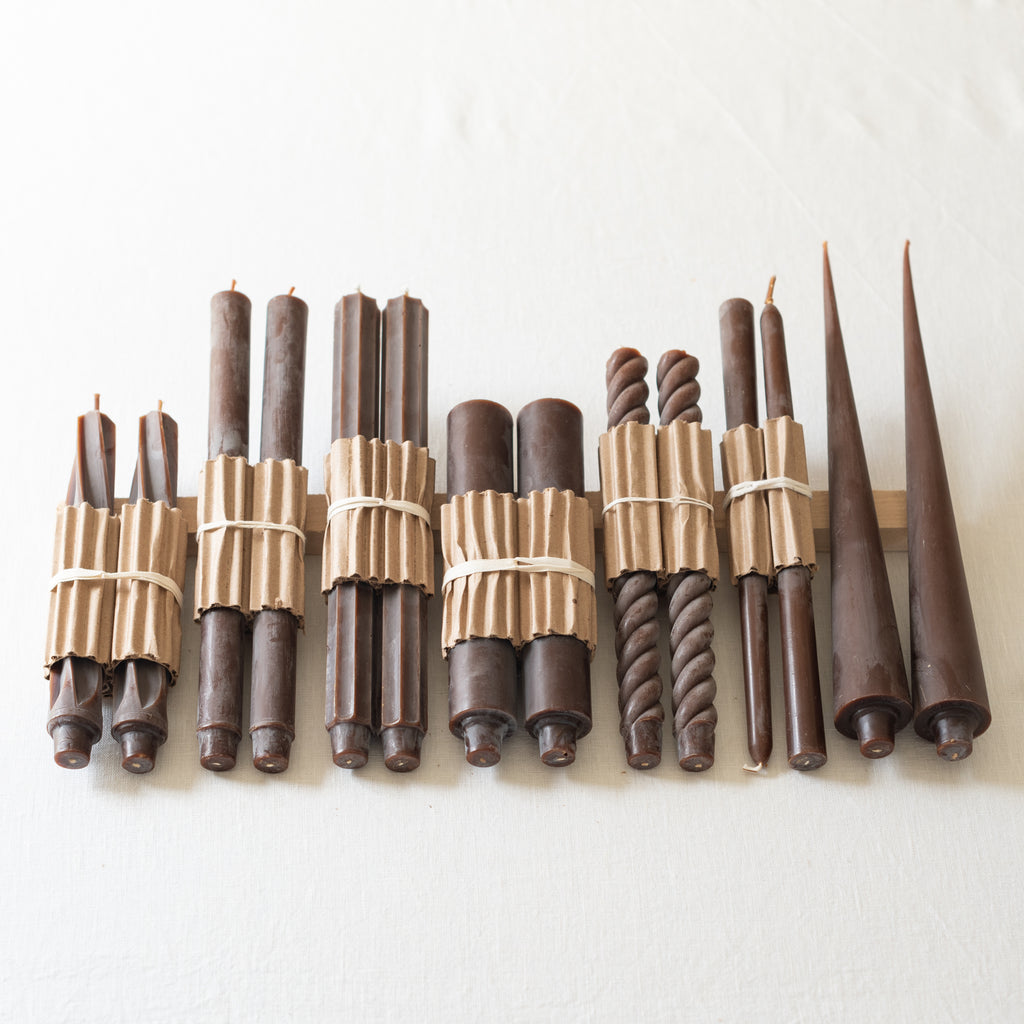 A selection of hand-poured taper candles in various shapes and sizes, all in a dark brown/espresso color, displayed against a neutral background.