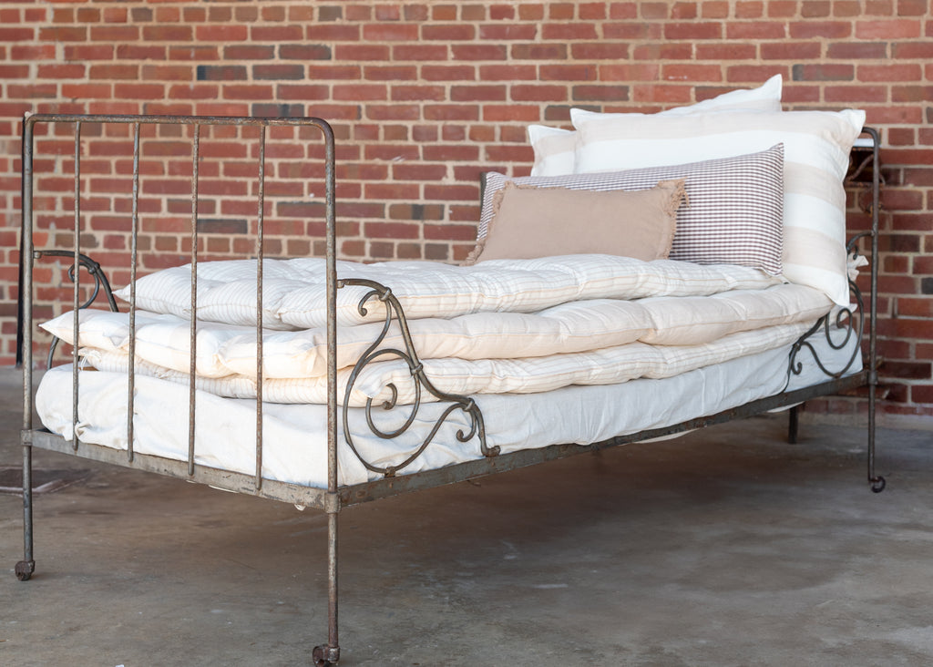 A white cotton mattress topper displayed on a bed with a metal frame, against a brick wall background.