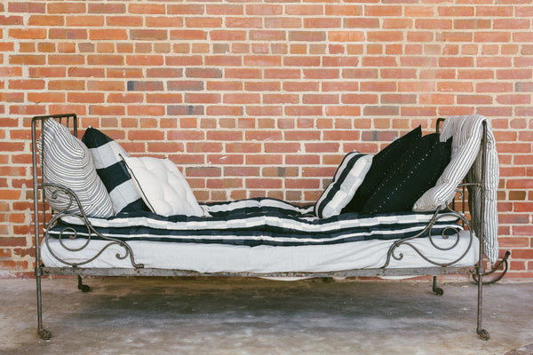 A striped cotton mattress topper displayed on a bed with a metal frame, against a brick wall background.