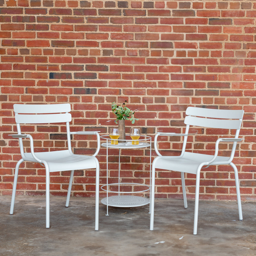 Two white Luxembourg armchairs, placed on either side of a round side table, against a brick wall background.